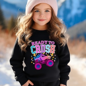 Pu&ograve; includere: Una felpa nera con la scritta "READY TO CRUSH" in lettere colorate e una grafica di un monster truck. Il monster truck &egrave; rosa, viola e blu con stelle e fulmini. La persona indossa un berretto rosa.
