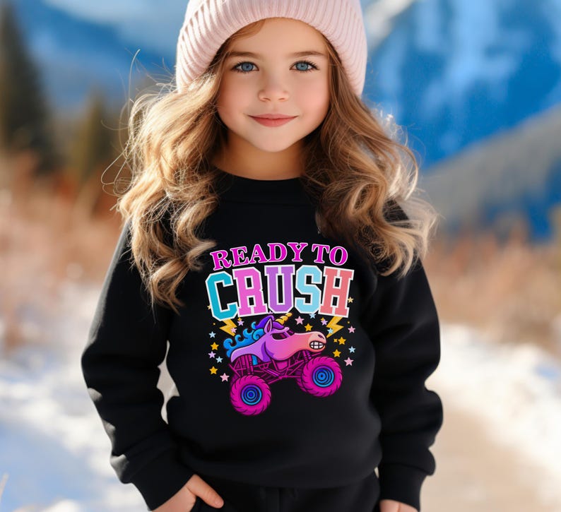 Pu&ograve; includere: Una felpa nera con la scritta "READY TO CRUSH" in lettere colorate e una grafica di un monster truck. Il monster truck &egrave; rosa, viola e blu con stelle e fulmini. La persona indossa un berretto rosa.