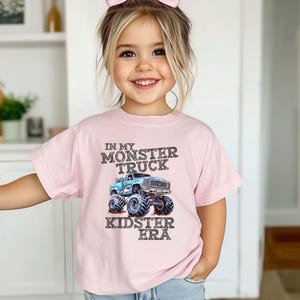 Puede incluir: Una camiseta rosa con un gr&aacute;fico de cami&oacute;n monstruo azul y el texto "In My Monster Truck Kidster Era".