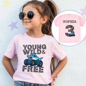 Op de afbeelding: Een lichtroze t-shirt met de tekst "YOUNG WILD & FREE" en een haai op monster truck wielen. Het shirt heeft ook een kleinere afbeelding van een roze shirt met de naam "SOPHIA" en het nummer "3".
