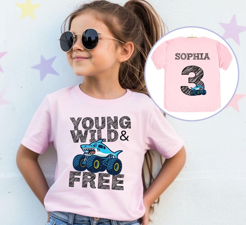 Op de afbeelding: Een lichtroze t-shirt met de tekst "YOUNG WILD & FREE" en een haai op monster truck wielen. Het shirt heeft ook een kleinere afbeelding van een roze shirt met de naam "SOPHIA" en het nummer "3".
