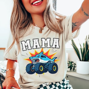 May include: Beige t-shirt with the word "MAMA" in a bold, outlined font. Below the text is a graphic of a blue shark monster truck with yellow and red accents. The shirt is a casual style.