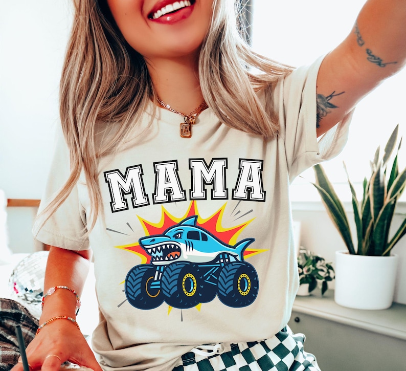 May include: Beige t-shirt with the word "MAMA" in a bold, outlined font. Below the text is a graphic of a blue shark monster truck with yellow and red accents. The shirt is a casual style.