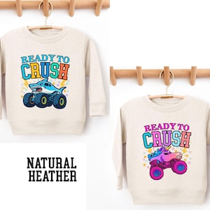 Pu&ograve; includere: Due felpe in heather naturale con la scritta "READY TO CRUSH". Una felpa presenta uno squalo su un monster truck, l'altra un unicorno su un monster truck. Il testo "NATURAL HEATHER" &egrave; in basso.