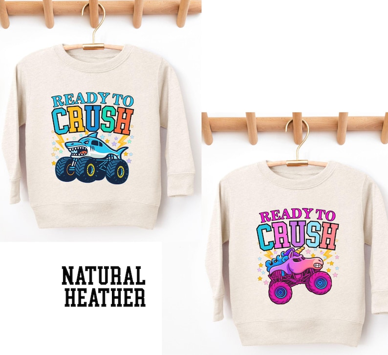 Pu&ograve; includere: Due felpe in heather naturale con la scritta "READY TO CRUSH". Una felpa presenta uno squalo su un monster truck, l'altra un unicorno su un monster truck. Il testo "NATURAL HEATHER" &egrave; in basso.