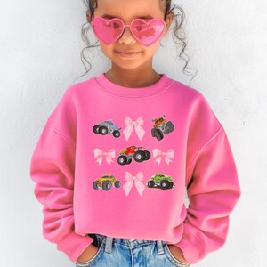 May include: A bright pink sweatshirt featuring colorful monster truck graphics and pink bow accents. The sweatshirt has a crew neck and long sleeves. The design includes a variety of monster trucks in different colors.