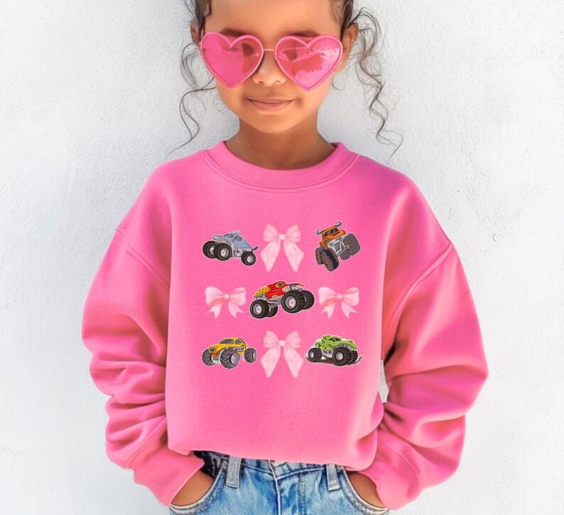 May include: A bright pink sweatshirt featuring colorful monster truck graphics and pink bow accents. The sweatshirt has a crew neck and long sleeves. The design includes a variety of monster trucks in different colors.