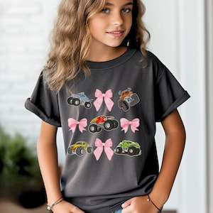 K&ouml;nnte beinhalten: Dunkelgraues T-Shirt mit bunten Monstertruck-Designs und rosa Schleifen. Die Trucks sind in verschiedenen Farben wie Blau, Orange, Rot, Gelb und Gr&uuml;n gehalten. Das T-Shirt hat einen l&auml;ssigen Stil.