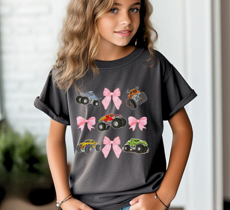 K&ouml;nnte beinhalten: Dunkelgraues T-Shirt mit bunten Monstertruck-Designs und rosa Schleifen. Die Trucks sind in verschiedenen Farben wie Blau, Orange, Rot, Gelb und Gr&uuml;n gehalten. Das T-Shirt hat einen l&auml;ssigen Stil.