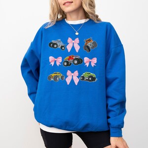 May include: A royal blue sweatshirt featuring embroidered monster trucks and pink bows. The design includes six different monster truck illustrations in various colors, paired with delicate pink bows. The sweatshirt is a comfortable, casual style.
