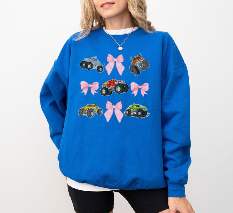 May include: A royal blue sweatshirt featuring embroidered monster trucks and pink bows. The design includes six different monster truck illustrations in various colors, paired with delicate pink bows. The sweatshirt is a comfortable, casual style.