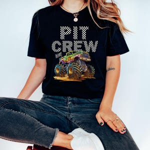 May include: Black t-shirt with the words "PIT CREW" in a checkered pattern above a colorful monster truck graphic. The monster truck is green, purple, and gold, with large tires. The shirt is worn by a person.