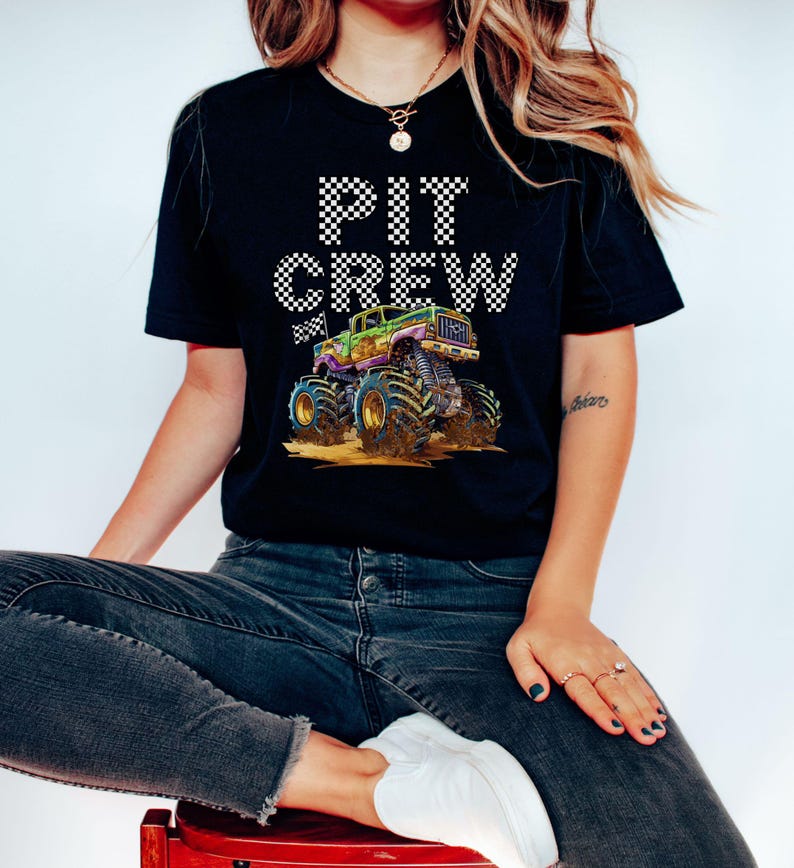 May include: Black t-shirt with the words "PIT CREW" in a checkered pattern above a colorful monster truck graphic. The monster truck is green, purple, and gold, with large tires. The shirt is worn by a person.
