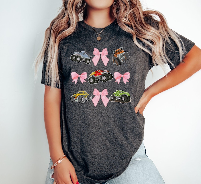 Pictured is a lady wearing a dark gray t-shirt featuring colorful monster Character truck graphics and pink bows. The design includes trucks in blue, orange, red, yellow, and green. A fun and playful design for fans of monster trucks.