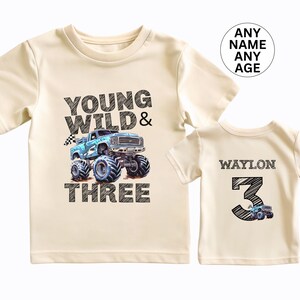 May include: Two cream-colored t-shirts. The front of the larger shirt has the text "YOUNG WILD & THREE" with a blue monster truck graphic. The smaller shirt has the name "WAYLON" and the number "3" with a monster truck graphic.