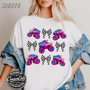 May include: White t-shirt featuring a repeating pattern of pink monster trucks with unicorn horns and black and white checkered bows. The word "WHITE" is at the top. Designed by Common Code.