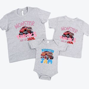 May include: Three gray t-shirts and a onesie with monster truck graphics. The shirts have the text "MONSTER TRUCKS ARE MY JAM" in pink. The onesie says "KIDSTER JAM" with a monster truck illustration.