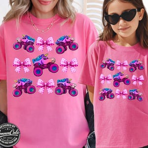 May include: Pink t-shirts with a repeating pattern of a unicorn monster truck and pink bows. The shirts are worn by a woman and a child. The text "DESIGNED BY Common Code EST. 2020 FOR YOU AND YOUR CREW" is visible.