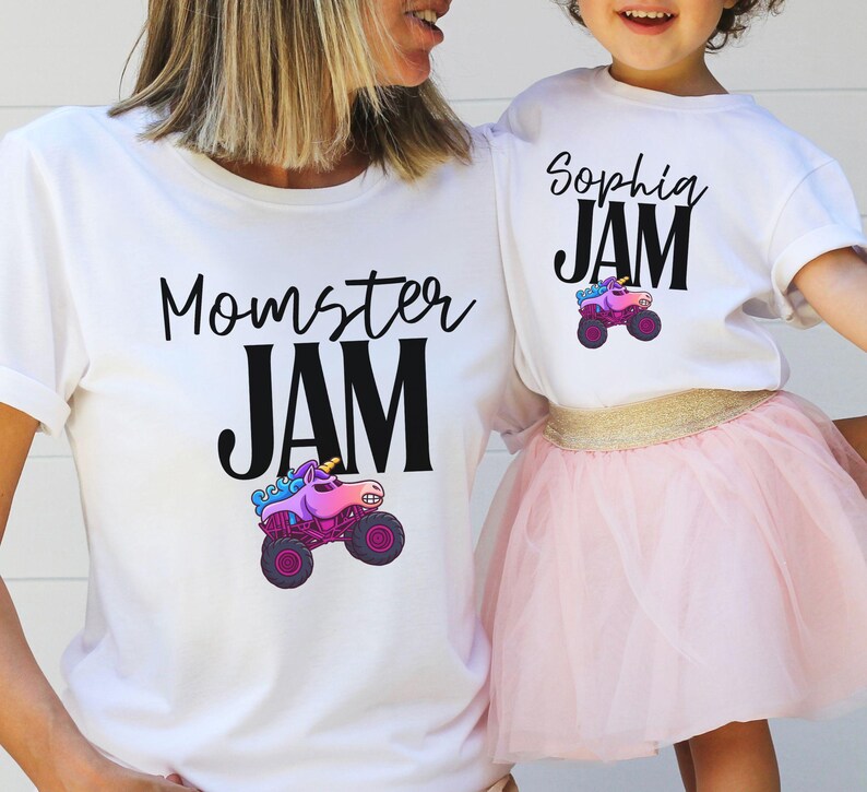May include: White t-shirts with matching designs. One shirt says "Momster JAM" and the other says "Sophia JAM". Both shirts feature a colorful monster truck with a unicorn design. The child is wearing a pink tutu.
