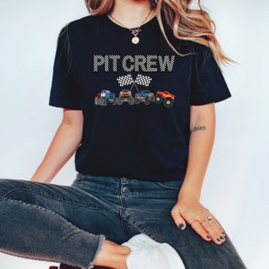 Puede incluir: Camiseta negra con el texto "PT CREW" en blanco con un dise&ntilde;o de bandera a cuadros. Cuatro monster trucks est&aacute;n alineados debajo del texto.