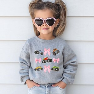 May include: A gray sweatshirt featuring embroidered monster trucks in various colors, including red, yellow, and green, interspersed with pink bows. The sweatshirt is worn by a young person wearing heart-shaped sunglasses.