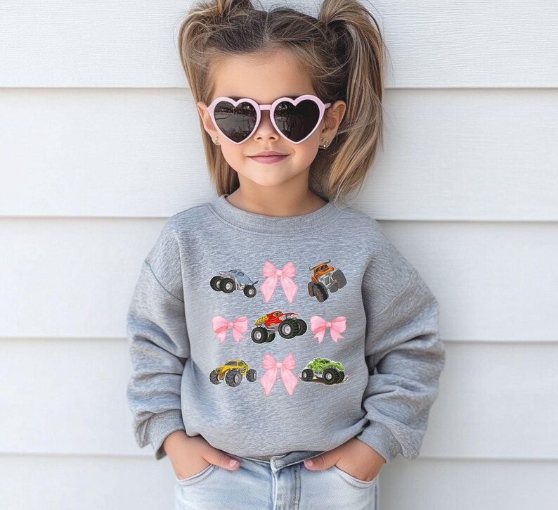 May include: A gray sweatshirt featuring embroidered monster trucks in various colors, including red, yellow, and green, interspersed with pink bows. The sweatshirt is worn by a young person wearing heart-shaped sunglasses.