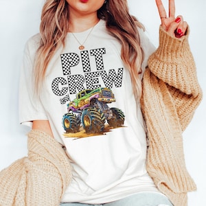 May include: Cream-colored t-shirt featuring the words "PIT CREW" in a checkered pattern above a colorful monster truck graphic. The monster truck has large tires and is in shades of green, purple, and yellow.