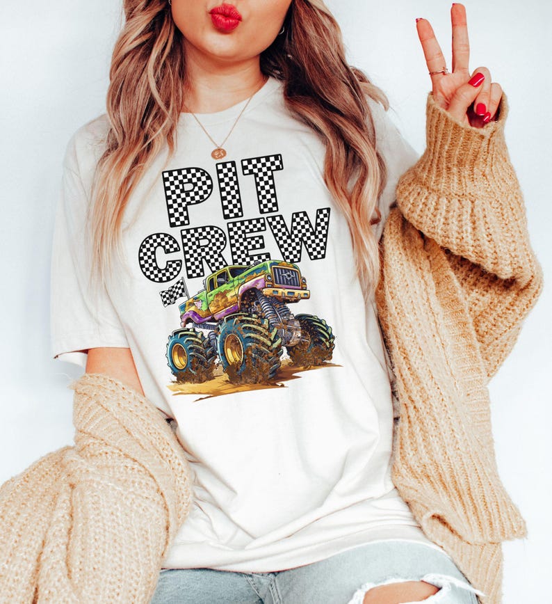 May include: Cream-colored t-shirt featuring the words "PIT CREW" in a checkered pattern above a colorful monster truck graphic. The monster truck has large tires and is in shades of green, purple, and yellow.