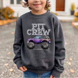 Puede incluir: Sudadera gris oscuro con las palabras "PIT CREW" en letras blancas. Un cami&oacute;n monstruo morado con neum&aacute;ticos negros grandes y una bandera a cuadros est&aacute; impreso en la parte delantera. La sudadera est&aacute; hecha de un material suave y c&oacute;modo.