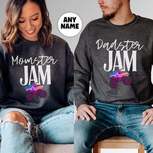 May include: Two dark gray sweatshirts with the text "Momster JAM" and "Dadster JAM" in white. Each sweatshirt features a colorful monster truck with a unicorn design. The sweatshirts are worn with blue jeans.