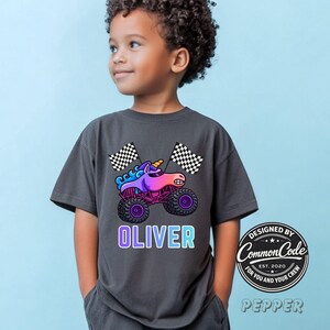 May include: Dark gray t-shirt featuring a colorful cartoon monster truck with a unicorn design, checkered flags, and the name "OLIVER" in a gradient font. The shirt also has a circular logo that says "Designed by Common Code".