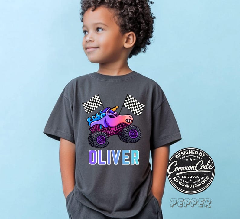 May include: Dark gray t-shirt featuring a colorful cartoon monster truck with a unicorn design, checkered flags, and the name "OLIVER" in a gradient font. The shirt also has a circular logo that says "Designed by Common Code".