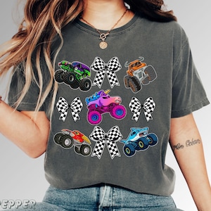 Comfort Colors t shirt in Pepper  race day ready with collage of croquette Black and White checkered bows and various monster truck jam trucks including Pink Sparkle Unicorn truck, Blue Shark Megalodon, Toro Bull, Green and Purple Digger, and Red Dragon Monster Truck. Very Vibrant and Fun Mom tee , matching mama and mini girls and boys monster truck party outfits. Ready for any Celebration or Gift for a Monster Truck lover. Adult and  Child kid sizes. Perfect for a teen gift or boy mama or dada
