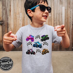 May include: A light gray t-shirt featuring colorful monster truck designs, including a unicorn, shark, and various other trucks. Each truck is paired with a checkered flag. The shirt also has the text "DESIGNED BY COMMON CODE EST. 2020 FOR YOU AND YOUR CREW".