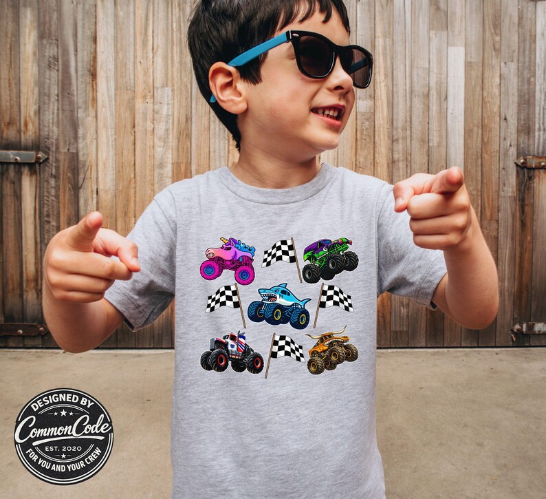 May include: A light gray t-shirt featuring colorful monster truck designs, including a unicorn, shark, and various other trucks. Each truck is paired with a checkered flag. The shirt also has the text "DESIGNED BY COMMON CODE EST. 2020 FOR YOU AND YOUR CREW".