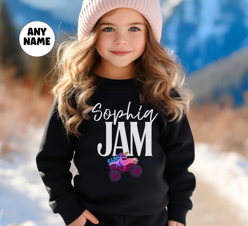 May include: Black sweatshirt with the name "Sophia" and the word "JAM" in white. A colorful unicorn monster truck graphic is below the word "JAM". The sweatshirt is worn by a person wearing a pink beanie.