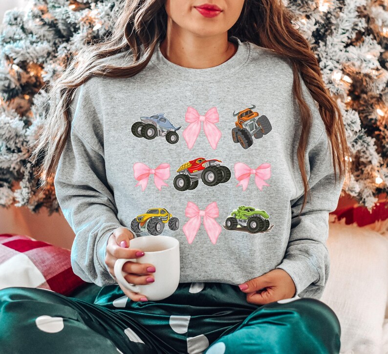 May include: A gray sweatshirt featuring colorful monster truck graphics and pink bows. The trucks are in various colors, including blue, orange, red, yellow, and green. The sweatshirt is a casual style.