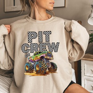 May include: A beige sweatshirt with a black and white checkered graphic that says "PIT CREW" and a cartoon monster truck with green, yellow, and black details.