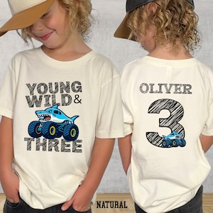 Op de afbeelding: Cr&egrave;mekleurig T-shirt met de tekst "YOUNG WILD & THREE" en een haai-thema monstertruck-afbeelding. De achterkant van het shirt heeft de naam "OLIVER" en het nummer "3" met een bijpassende truck-afbeelding. Het woord "NATURAL" staat onderaan.