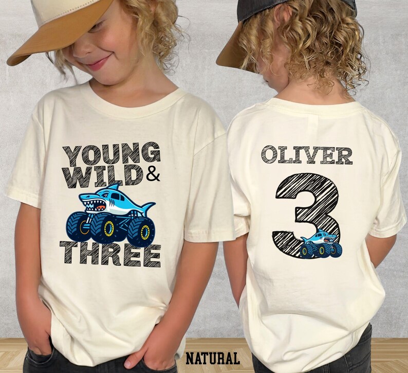 Op de afbeelding: Cr&egrave;mekleurig T-shirt met de tekst "YOUNG WILD & THREE" en een haai-thema monstertruck-afbeelding. De achterkant van het shirt heeft de naam "OLIVER" en het nummer "3" met een bijpassende truck-afbeelding. Het woord "NATURAL" staat onderaan.