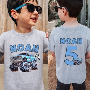 Pictured is a young boy wearing a light gray T-shirt with custom name Noah on front with a blue monster truck with shark theme and checkered flag. The back of the shirt features "NOAH" and the number "5" in blue. A small monster truck graphic is below the number.