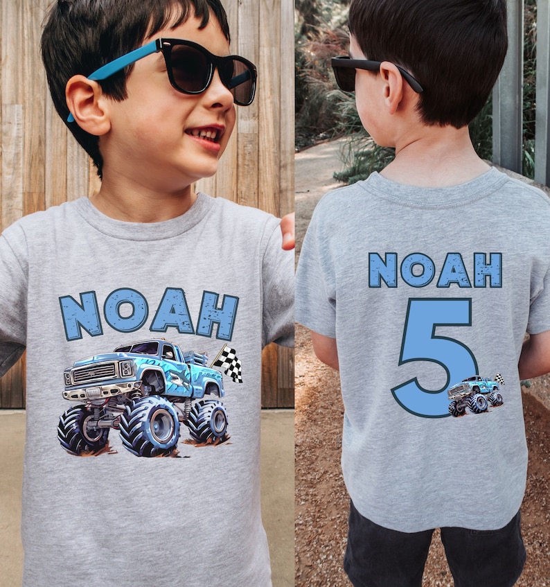 Pictured is a young boy wearing a light gray T-shirt with custom name Noah on front with a blue monster truck with shark theme and checkered flag. The back of the shirt features "NOAH" and the number "5" in blue. A small monster truck graphic is below the number.