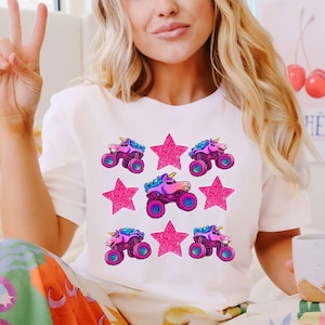 May include: White t-shirt featuring a repeating pattern of pink monster trucks with unicorn horns and pink glitter stars. The monster trucks have blue, purple, and pink accents. The shirt is a casual, fun design.