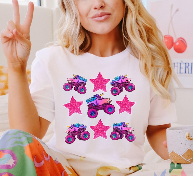 May include: White t-shirt featuring a repeating pattern of pink monster trucks with unicorn horns and pink glitter stars. The monster trucks have blue, purple, and pink accents. The shirt is a casual, fun design.