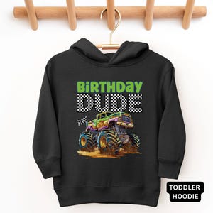 May include: Black toddler hoodie with the words "BIRTHDAY DUDE" in green and checkered text. A colorful monster truck graphic is featured below the text. The hoodie is hanging on a wooden hanger.
