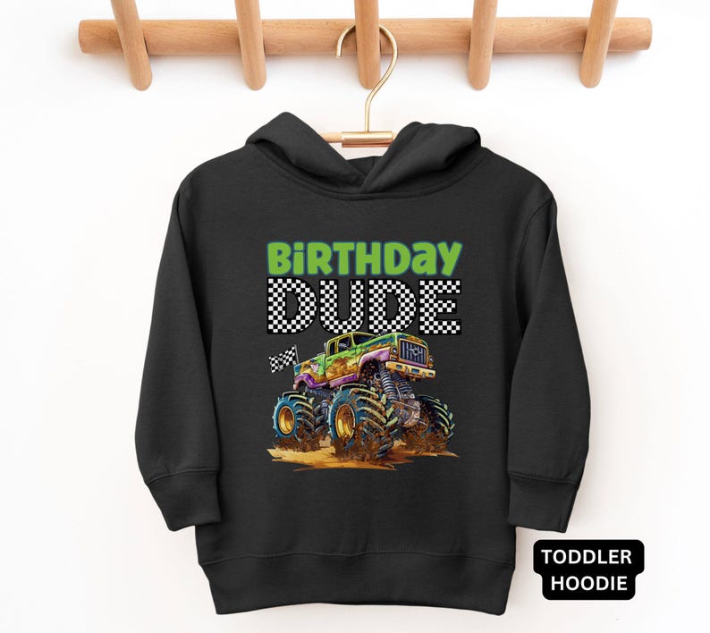 May include: Black toddler hoodie with the words "BIRTHDAY DUDE" in green and checkered text. A colorful monster truck graphic is featured below the text. The hoodie is hanging on a wooden hanger.