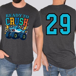 Dad's dark gray t-shirt with a shark monster truck graphic and the text "READY TO CRUSH" on the front. The back of the shirt features the number "29" in turquoise with a black outline. The shirt is made of a soft material.