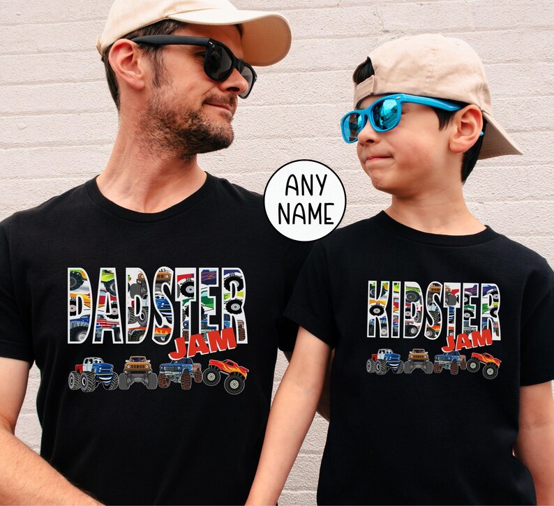father son models are wearing matching black t-shirts that are monster truck themed. "pattern letters that say "Dadster Jam" and "Kidster Jam". Each top has a row of 4 Monster Trucks underneath the letters.