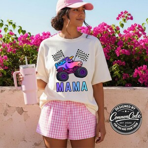May include: Cream-colored t-shirt featuring a colorful unicorn monster truck design with the word "MAMA" in gradient letters. The shirt is paired with pink and white checkered shorts. A pink tumbler with a straw is held in the left hand.