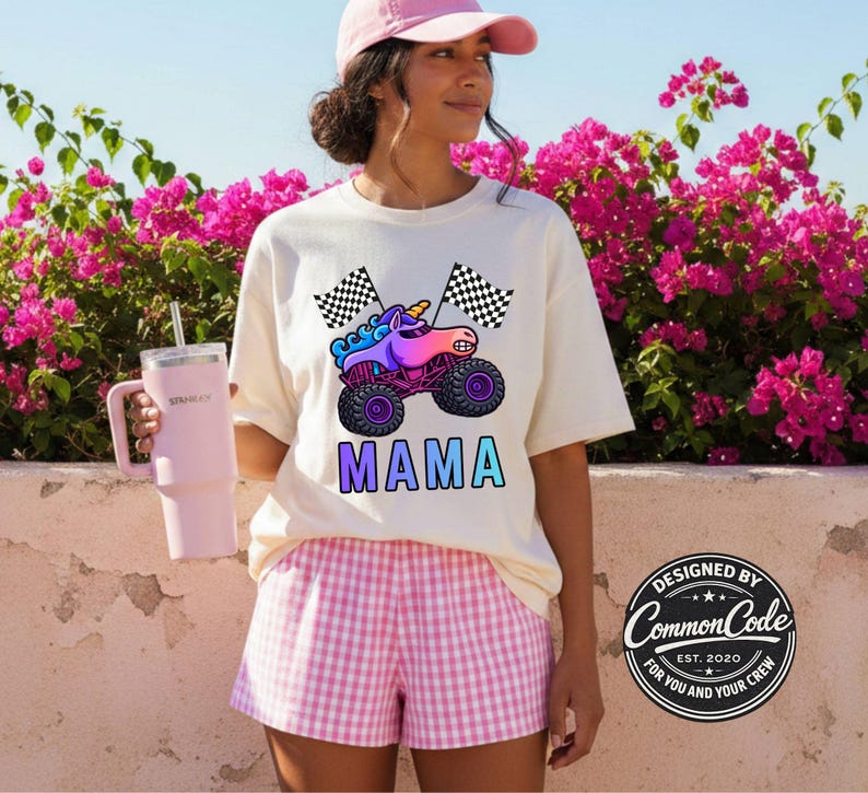 May include: Cream-colored t-shirt featuring a colorful unicorn monster truck design with the word "MAMA" in gradient letters. The shirt is paired with pink and white checkered shorts. A pink tumbler with a straw is held in the left hand.